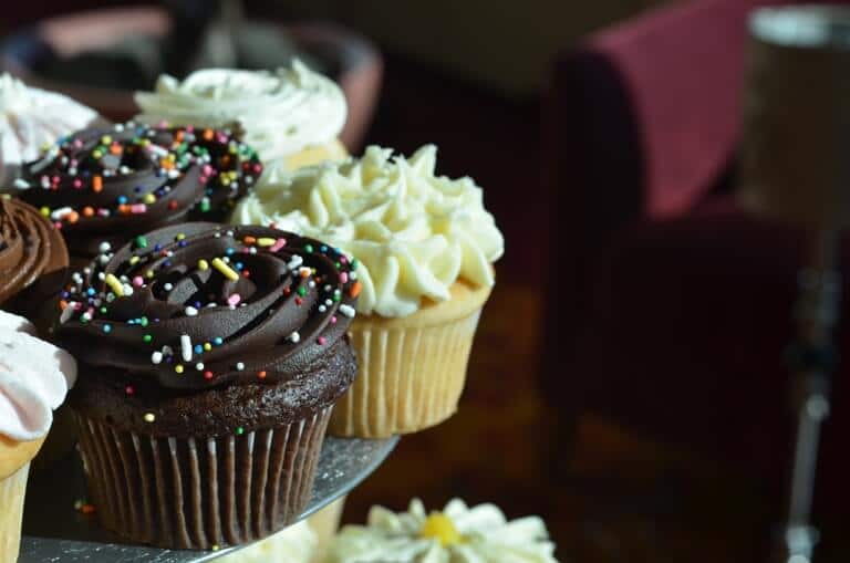 A close-up of different colored cupcakes with sprinkles on a table, a red chair in the background