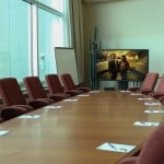 Papers and pens on a meeting table surrounded by red chairs, a woman and a man on the screen, and a white board in the background.