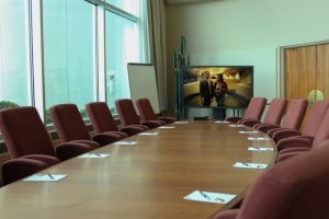 Papers and pens on a meeting table surrounded by red chairs, a woman and a man on the screen, and a white board in the background.