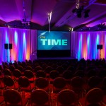 A distant view of a projector screen saying "It's Time", red chairs arranged ordery in an empty hall with colorful lights