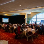 A group of people attending a seminar, gathered around round tables, a projector screen in the background