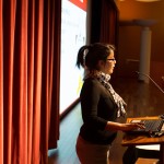 A woman speaker giving a speech behind a podium next to a projector screen