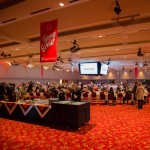 A large, crowded event venue, with long tables and chairs, a buffet in close-up, a woman grabbing drinks