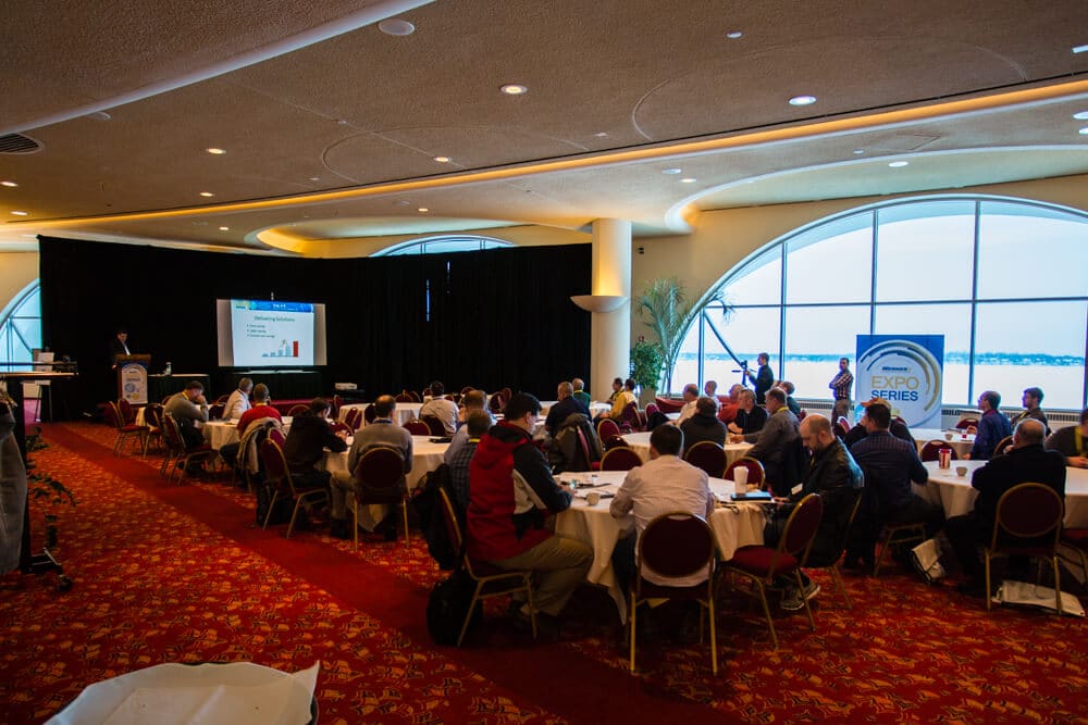 A meeting at the Grand Terrace with people gathered around tables, a speaker behind the podium, and a presentation on the screen