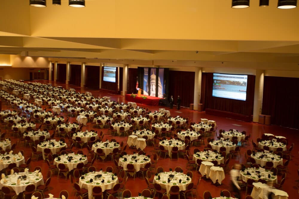 A large conference hall with dozens of round tables and chairs, three big screens and two men standing next to a platform.