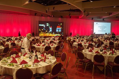 2015-Chamber-Dinner-HR-3575 A venue hall with round tables and two big screens, as staff works to set up the event.