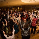 A group of people are dancing in formal outfits, their hands are up in the air in a venue