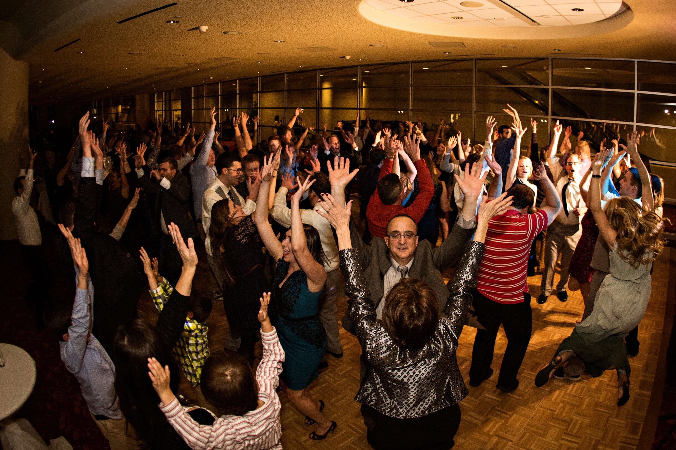A group of people are dancing in formal outfits, their hands are up in the air in a venue