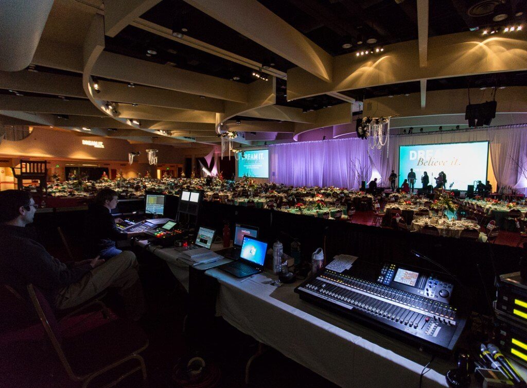 A technical crew working with computers and sound system at the back of a venue, as a band rehearses on the stage in the background