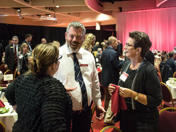 A man and two women talking during an event, with people and empty tables in the background