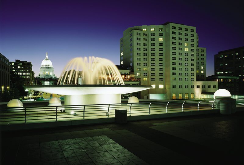 An ornamental pool lit up, city buildings and the Wisconsin State Capitol in the background at night.