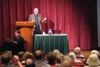 A speaker talking on stage behind a podium, a table with a green tablecloth in front of him, red curtains behind, audience watching