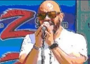 A man wearing sunglasses and a white shirt passionately sings into a microphone. A colorful backdrop with a large "Z" logo is in the background.