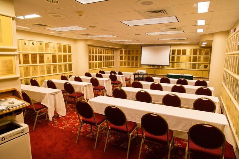 Nine long rectangular tables arranged in rows with red chairs, a white projector screen, and frames on the wall