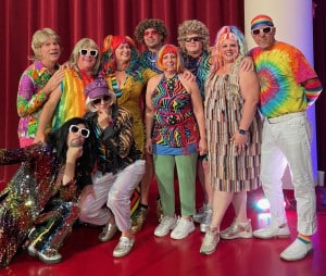 A group of people wearing vibrant, retro 70s outfits, including tie-dye and sequins, pose joyfully in front of a red curtain, exuding a playful, celebratory vibe.
