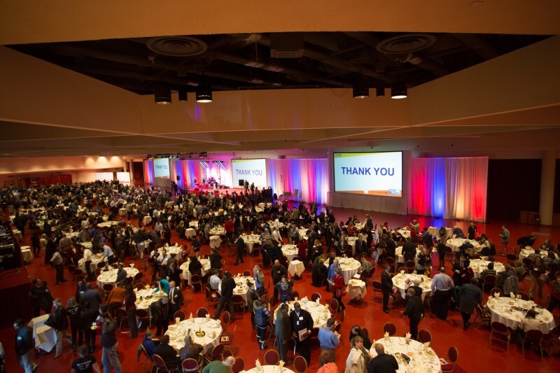 A distant view of a large event room with round tables and three big presentation screens