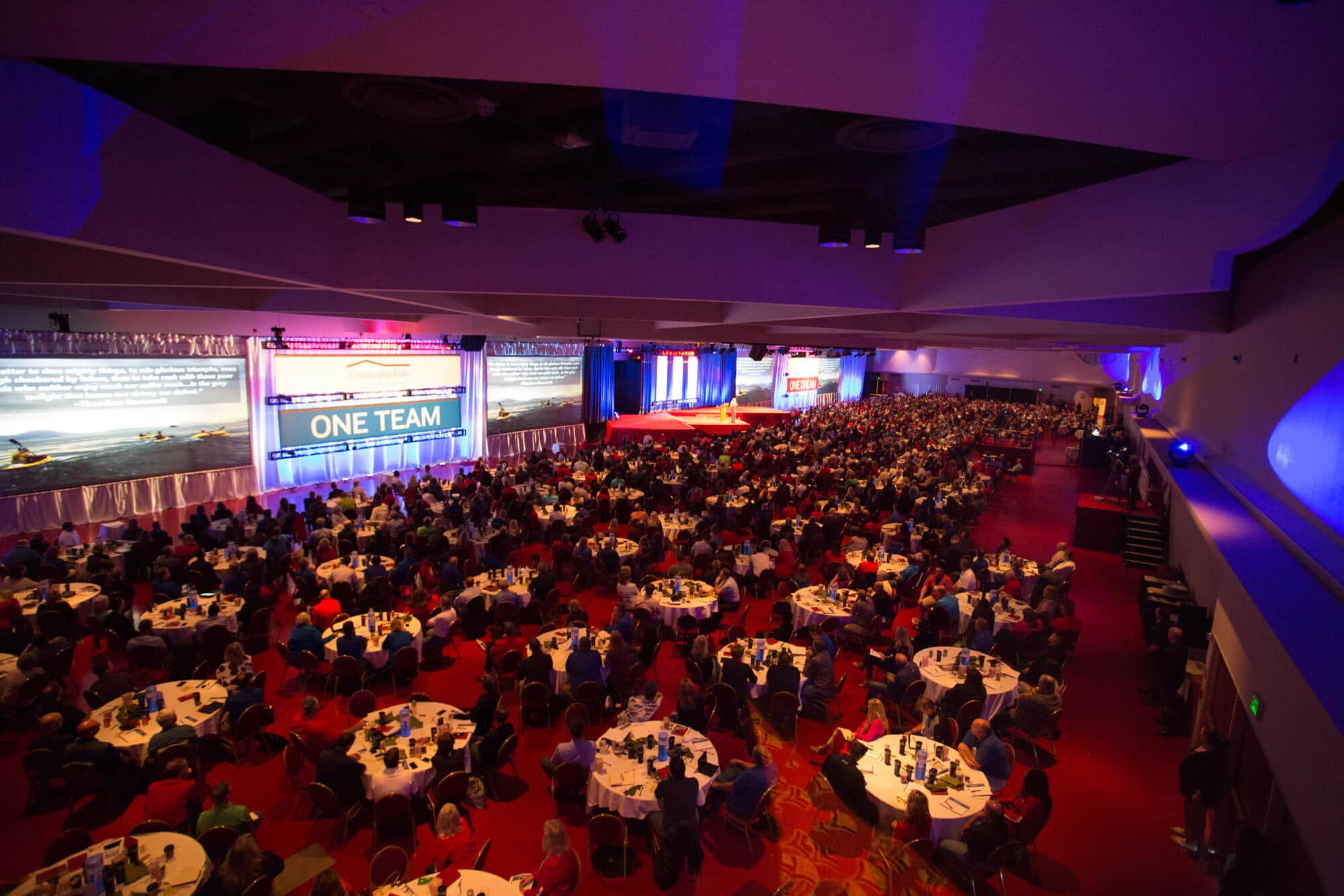 A large conference hall with dimmed lights, dozens of round tables, people gathered around, four big projector screens