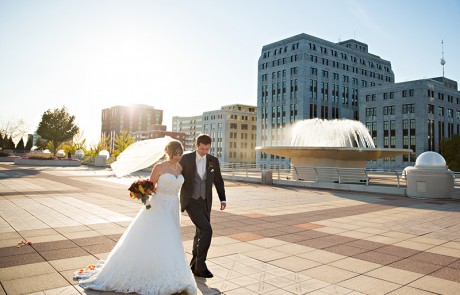 A newlywed couple walking together outside of Monona Terrace, buildings and a big ornamental pool in the background