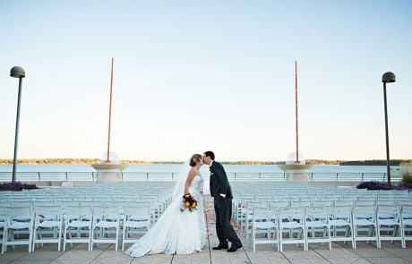 Bride and groom kissing by the lake, empty chairs and tall lamps in the background