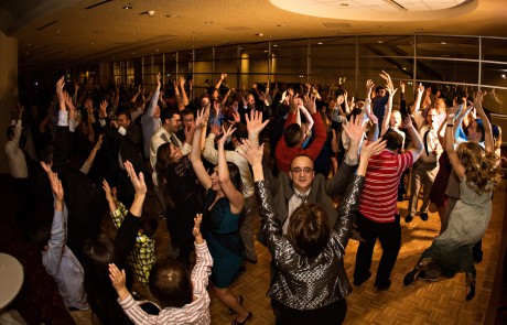 A group of people are dancing in formal outfits, their hands are up in the air in a venue