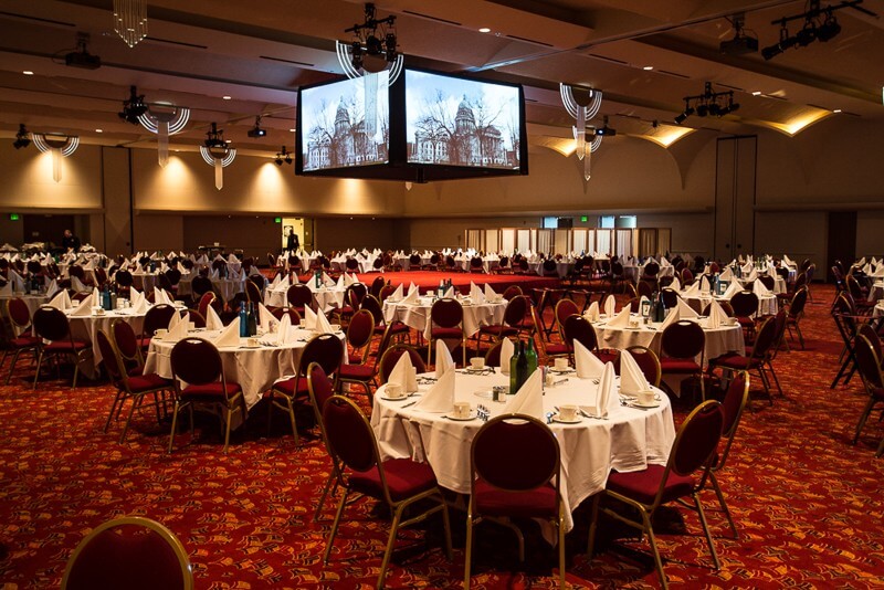 A dining hall with round tables and presentation screens on the ceiling.