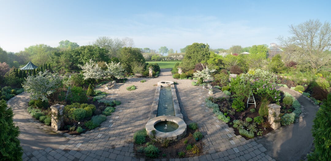 Aerial shot of a garden with a long ornamental pool in the middle, green trees and landscape plants, a green grass field in the background