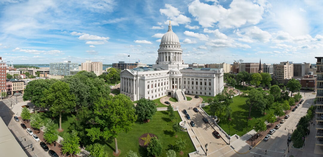 The Wisconsin State Capitol building among trees in daytime, city buildings in the background with a distant view of the lake.