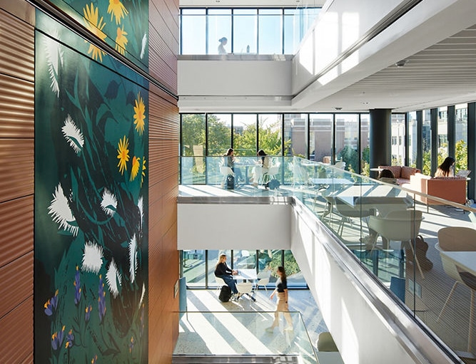 Bright, modern atrium with glass railings and skylight. Large floral mural adds vibrant colors. People work and relax, creating a serene atmosphere.
