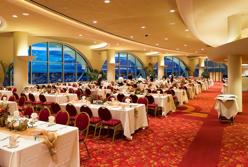 Decorated rectangular tables with red chairs in a venue with red carpet and big windows, staff working in the background