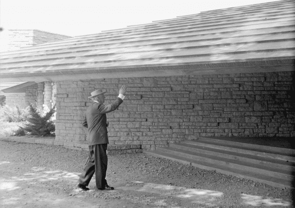 An old black-and-white photo of Frank Llyod Wright with a hat, pointing towards the low roofline at the Unitarian Meeting House in Madison, WI