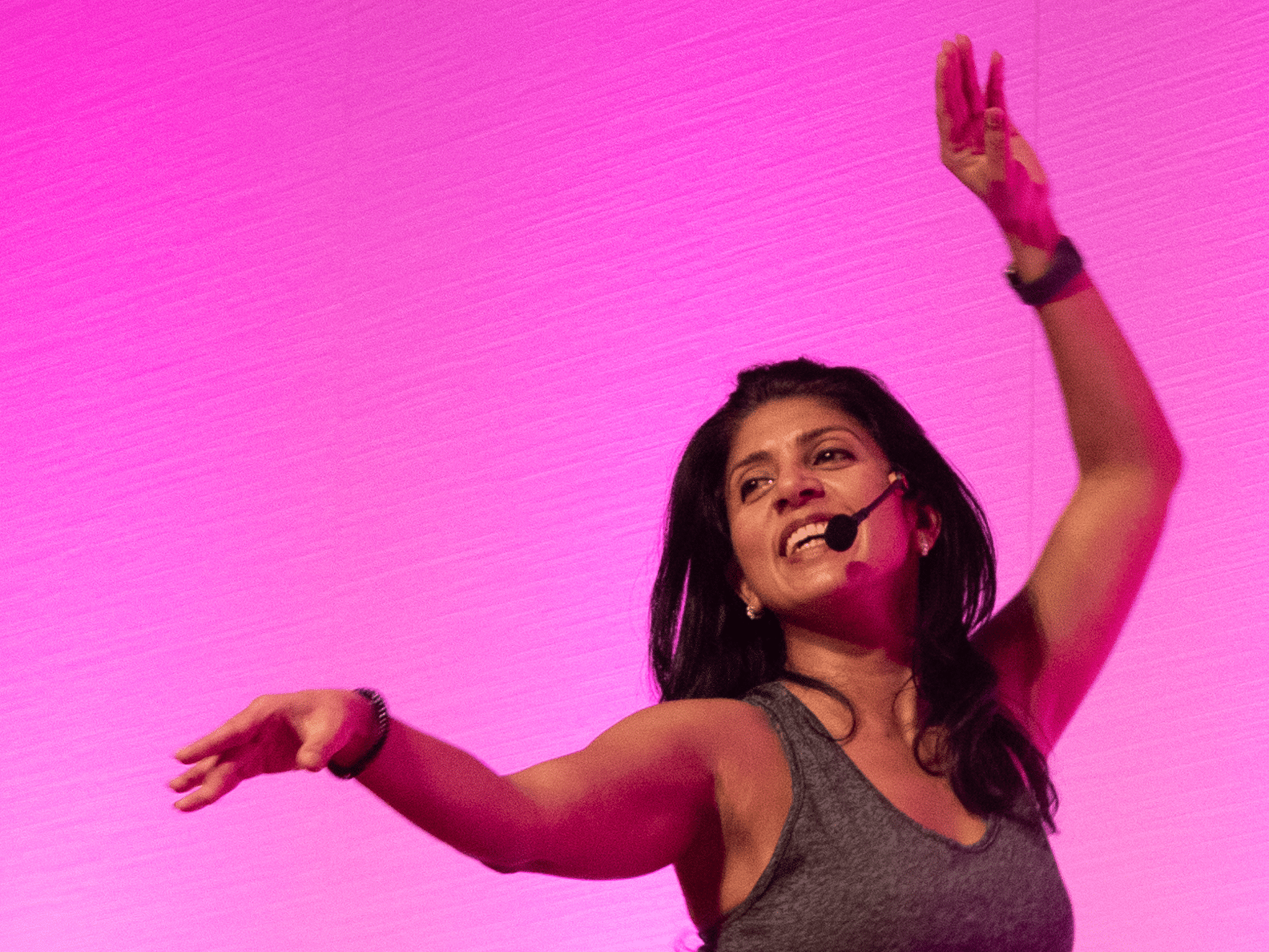 A woman joyfully dances with arms raised against a vibrant pink background, wearing a headset microphone and gray tank top, exuding energy and enthusiasm.