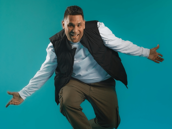 Man in a casual outfit is energetically jumping against a teal background, wearing a black vest and khaki pants, smiling with open arms.