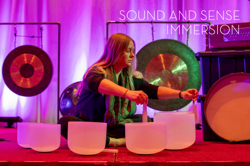 A woman sits cross-legged, playing crystal singing bowls on a red carpet. Behind her, gongs hang against a purple-lit backdrop. Text: "Sound and Sense Immersion."