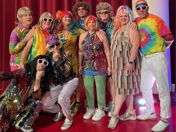 The band Vo5. A group of people in vibrant, colorful retro outfits, including tie-dye and sequins, pose joyfully against a red curtain backdrop, conveying fun and celebration.