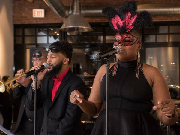 Soul 2 the Bone. Masquerade-themed musical performance with two vocalists wearing elegant masks, one in a red feathered mask, singing passionately into microphones. A trumpet player is in the background.