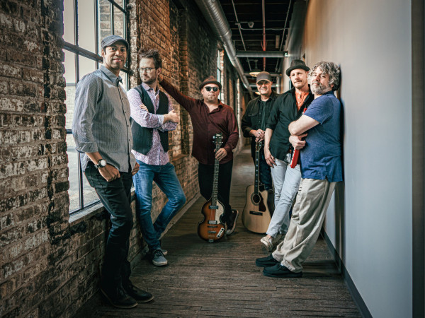 Love Monkeys. Six men casually pose in a narrow brick hallway with guitars. Their relaxed, confident expressions suggest a musical band vibe.