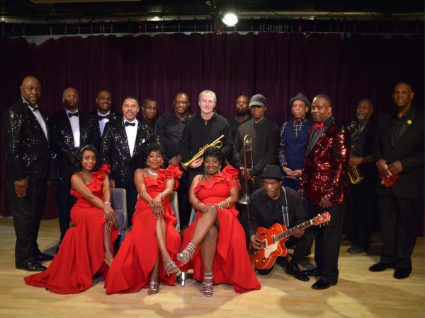 SOS Motown Revue. A jazz band group photo on stage features three women in elegant red dresses seated in front. Behind them, men in formal and glittering attire stand, some holding instruments, exuding a celebratory, vibrant atmosphere.
