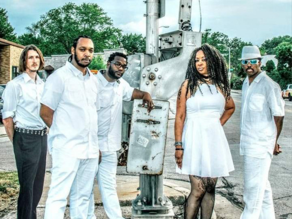 V-Funk. Five people in white attire stand confidently around an industrial device on a street, exuding a cool, relaxed vibe. Background shows trees and buildings.