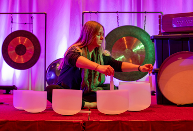 A woman sits on a red stage surrounded by crystal singing bowls and gongs, lit by soft pink and purple lighting. The scene conveys a calm, meditative atmosphere.