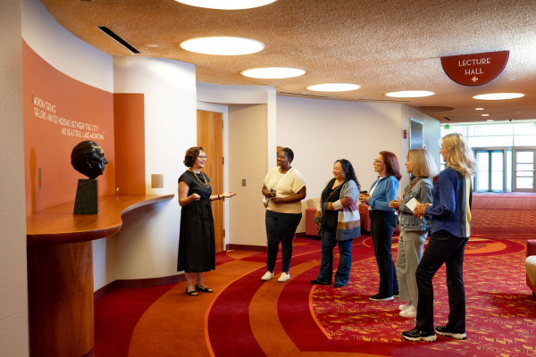 A smiling man gestures towards a modern building with large windows while leading a tour group outside. The scene is bright and relaxed.