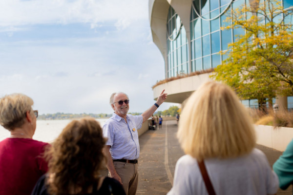 A smiling man gestures towards a modern building with large windows while leading a tour group outside. The scene is bright and relaxed.