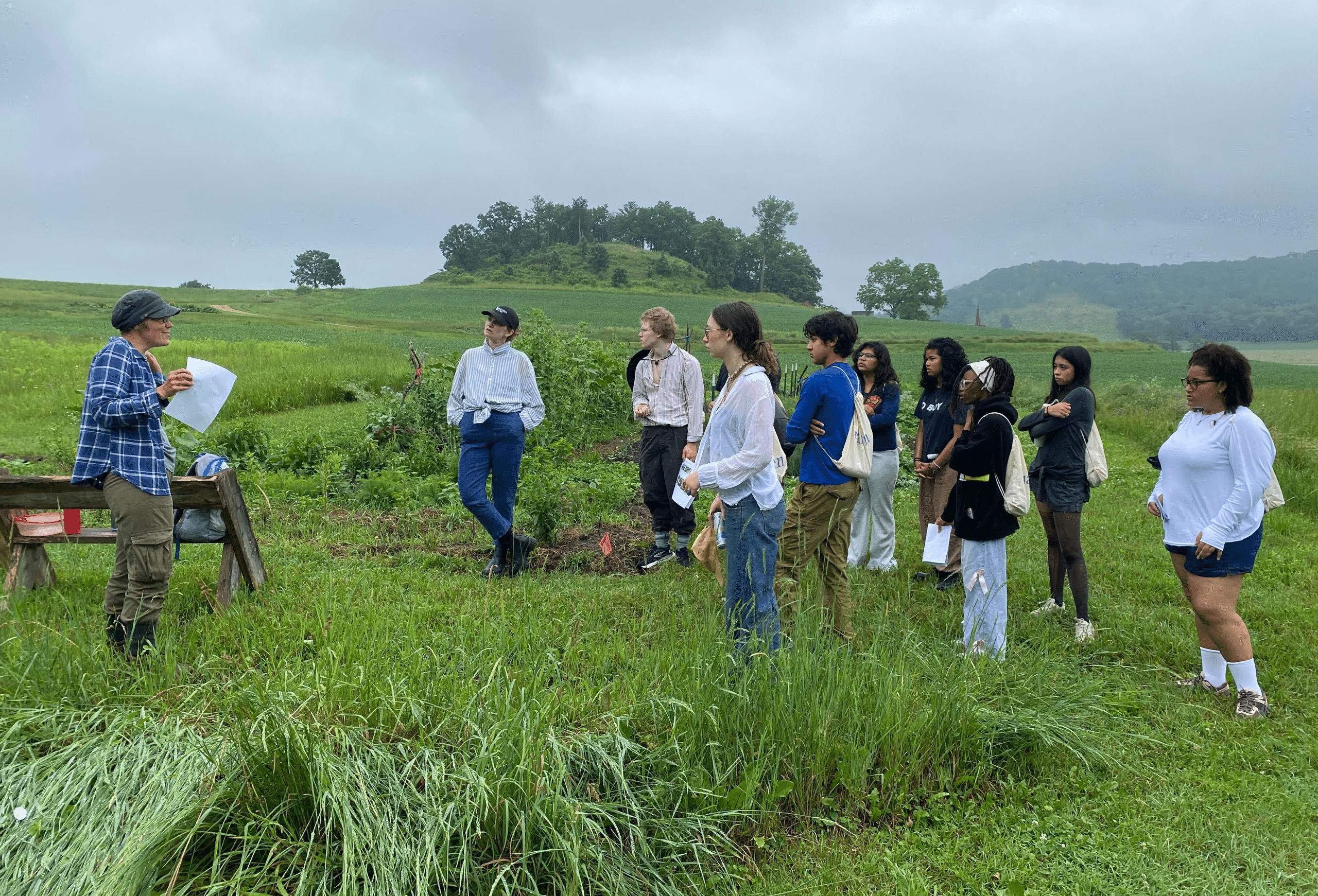 A group of people listen to a presenter outdoors in a lush, green field under a cloudy sky. The atmosphere is calm and attentive.