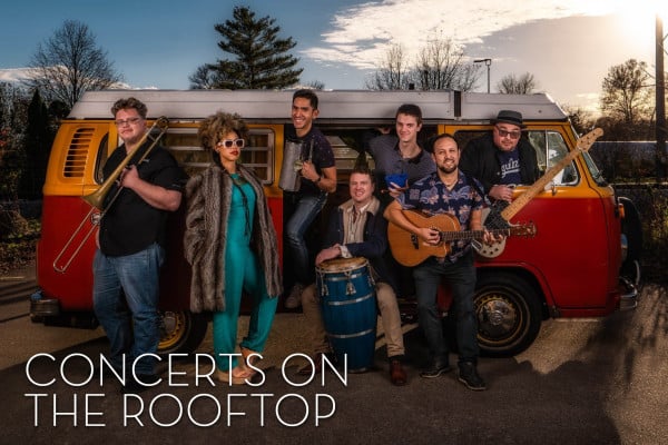 A diverse band gathers in front of a vintage red and yellow van, holding instruments and smiling warmly. Text reads "Concerts on the Rooftop."