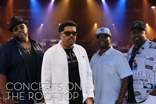 Four men in casual attire and sunglasses stand confidently on a rooftop stage, with colorful lights behind them. Text reads, “Concerts on the Rooftop.”