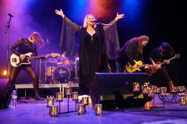 A band performs energetically on stage, with a female lead singer in black flowing sleeves singing passionately. Colorful lights illuminate the scene.