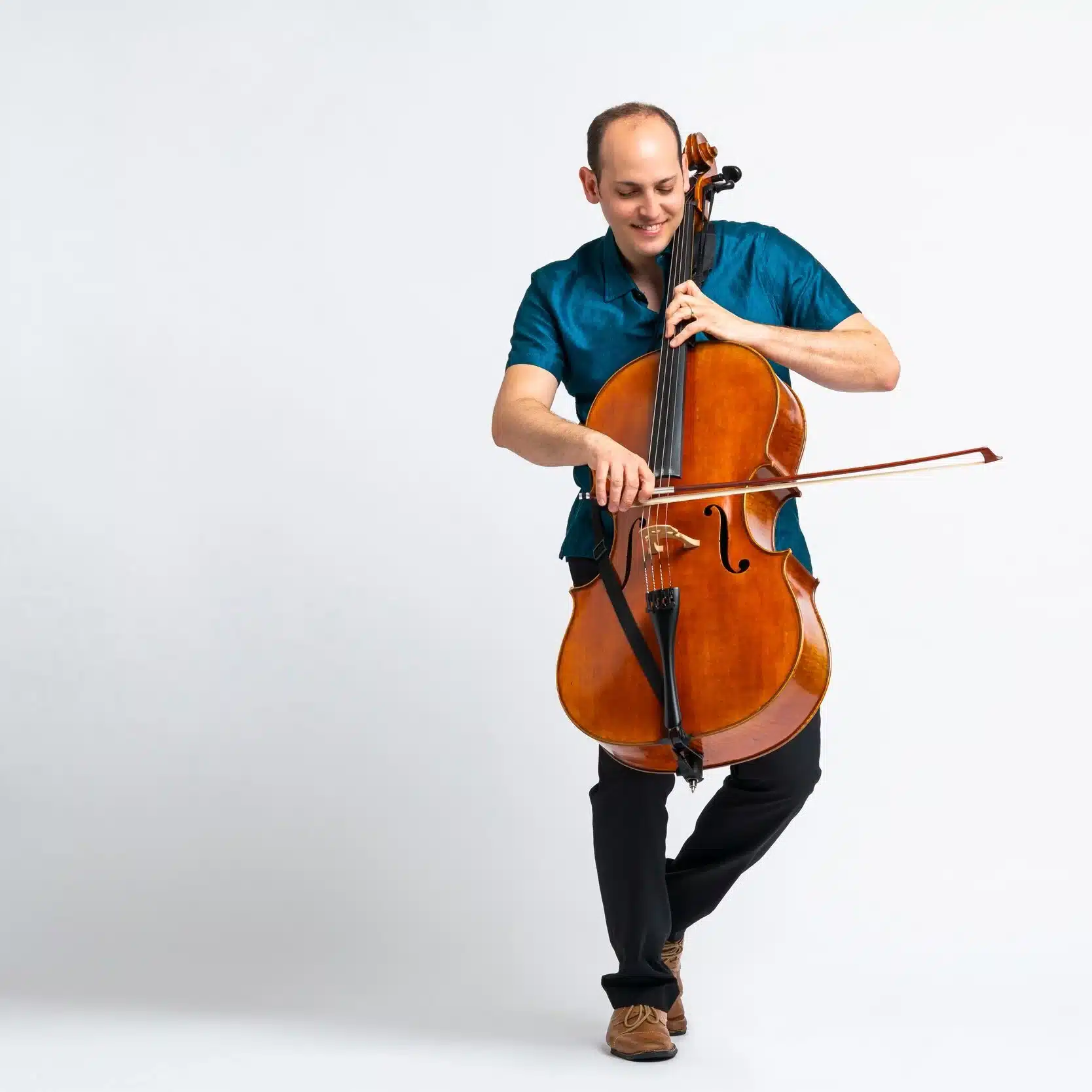 Mike Block in a teal shirt and black pants joyfully plays a cello. He stands on one leg against a plain white background, conveying passion and concentration.