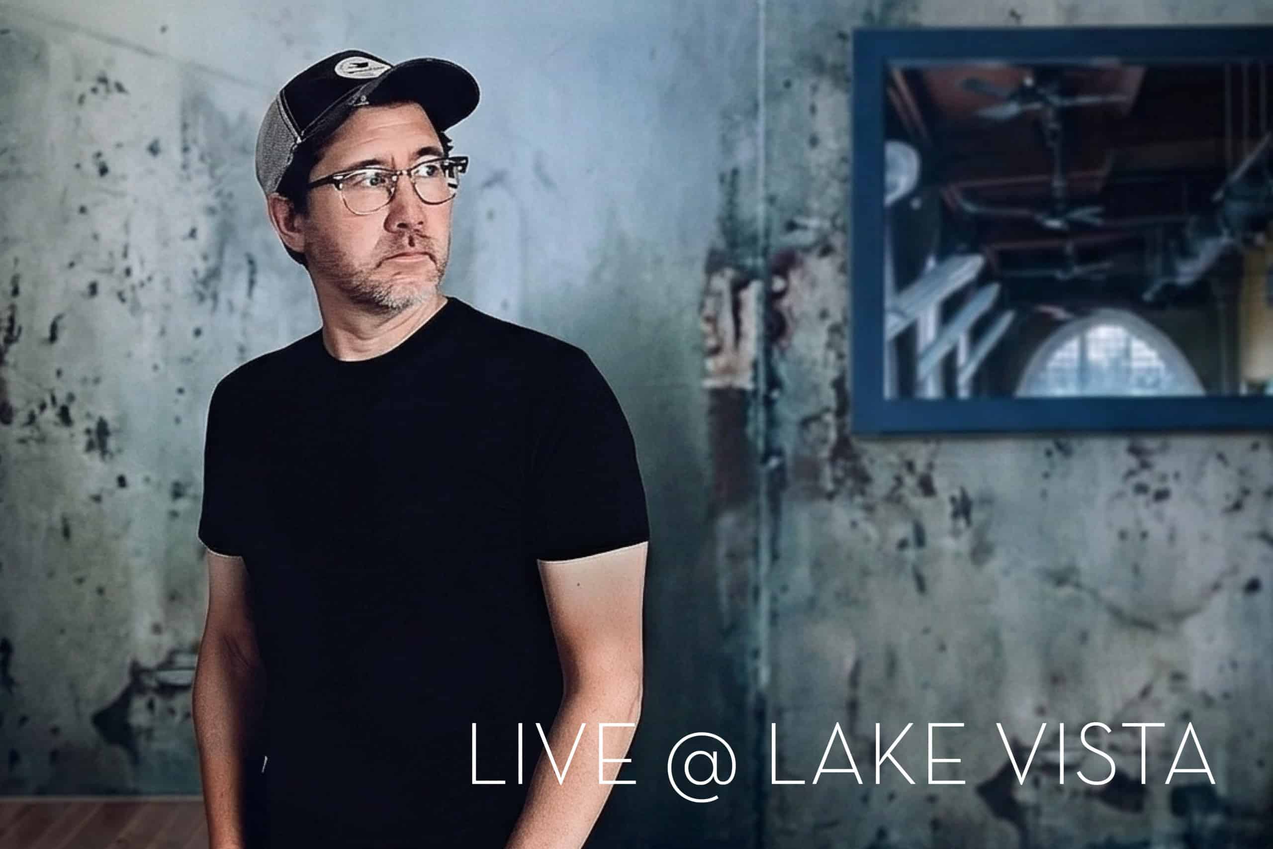 Jim White stands in front of a worn, textured wall with a window reflecting an industrial interior. Text reads “LIVE @ LAKE VISTA.”