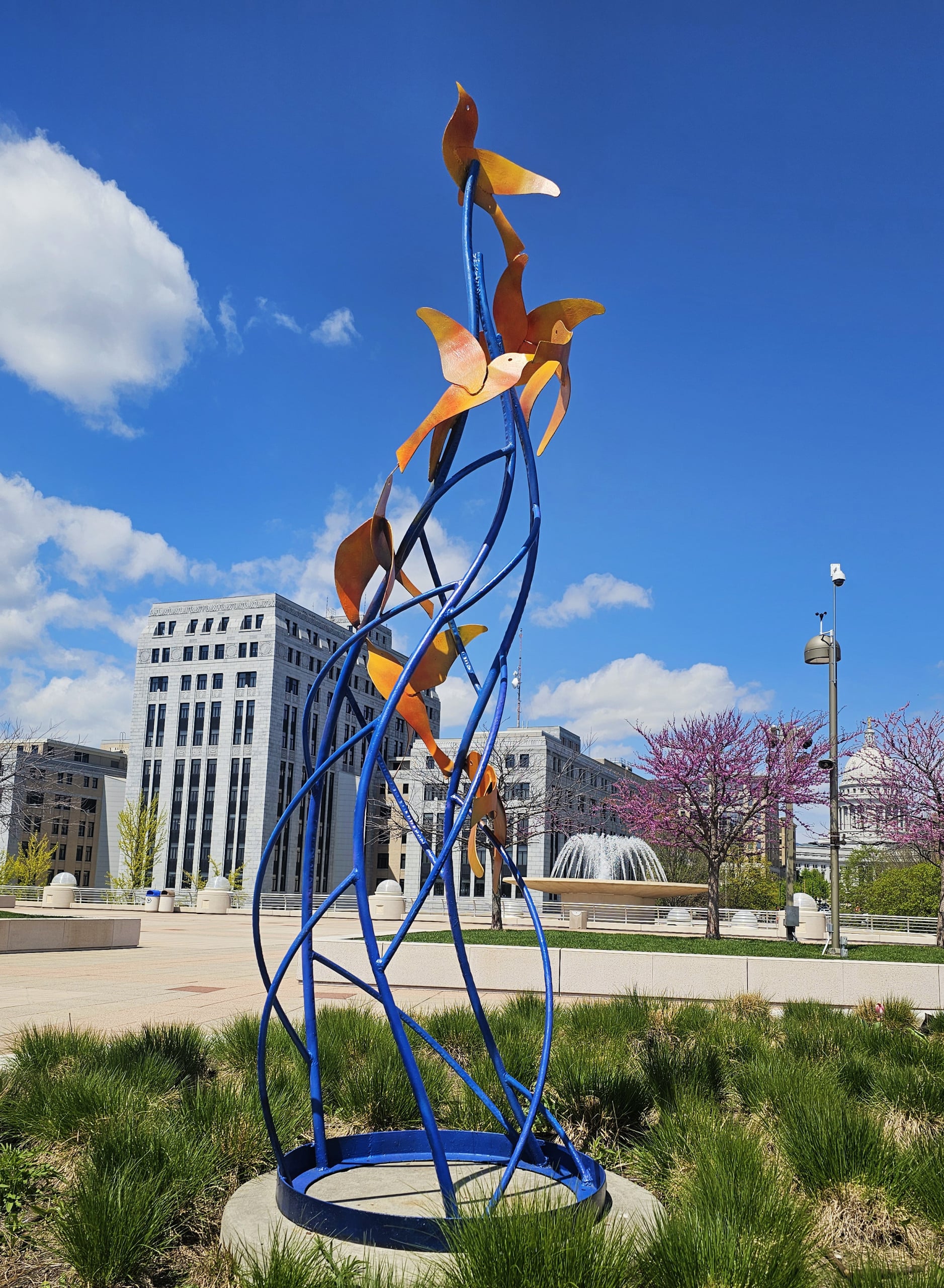 A vibrant outdoor sculpture with twisting blue metal and orange shapes stands amid greenery. Buildings and a fountain appear under a bright blue sky.