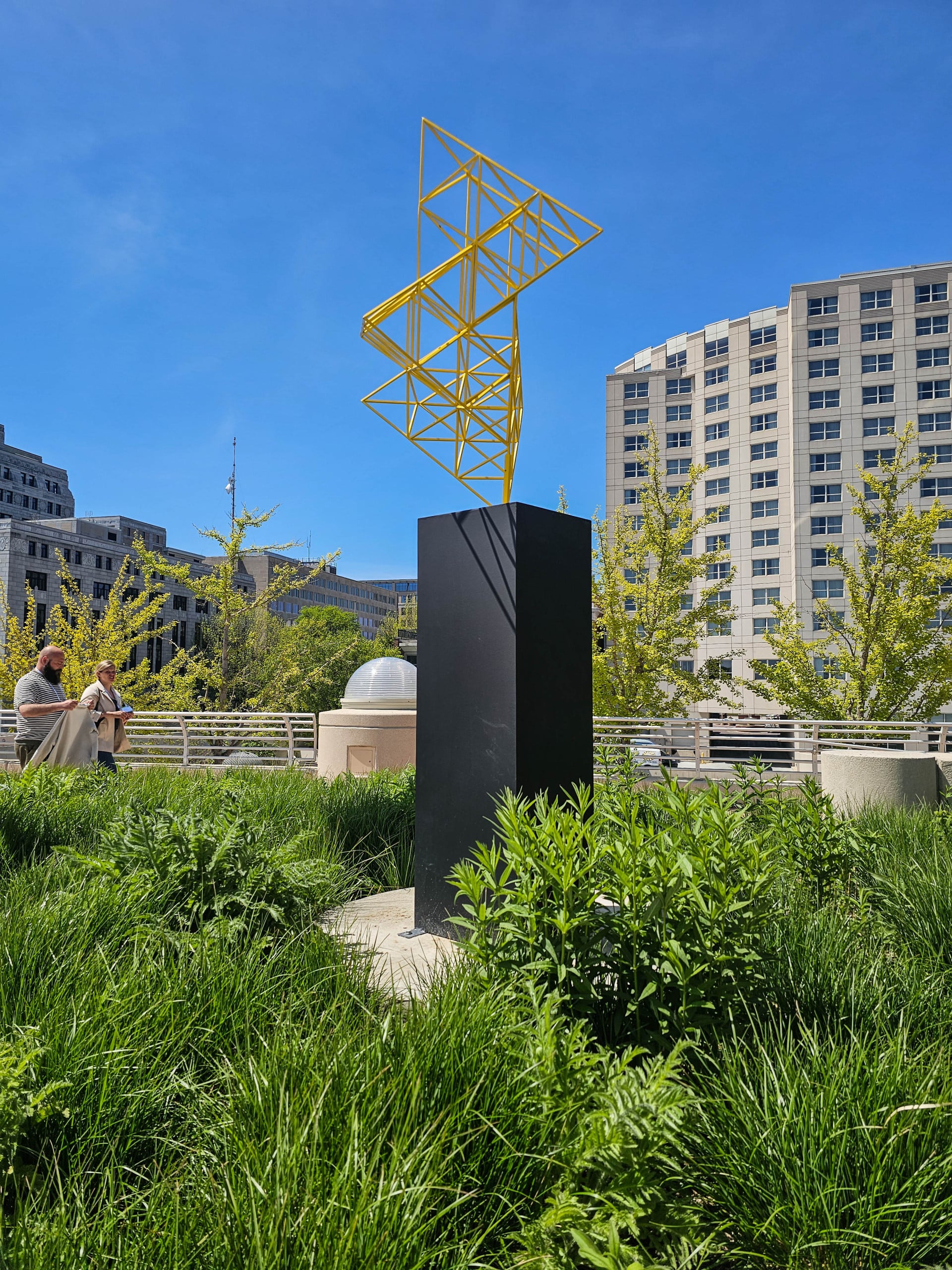 Bright yellow geometric metal sculpture on a black pedestal outside a modern building with large windows and neutral tones, creating a bold contrast.