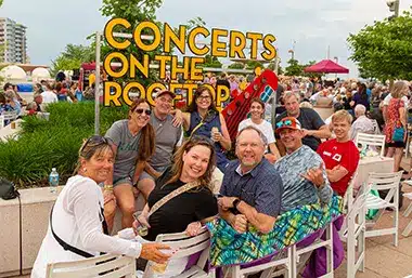 CORJUNE1-6187.png A group of people seated outdoors at "Concerts on the Rooftop," smiling and relaxed. Bright sign and crowd create a lively, festive atmosphere.